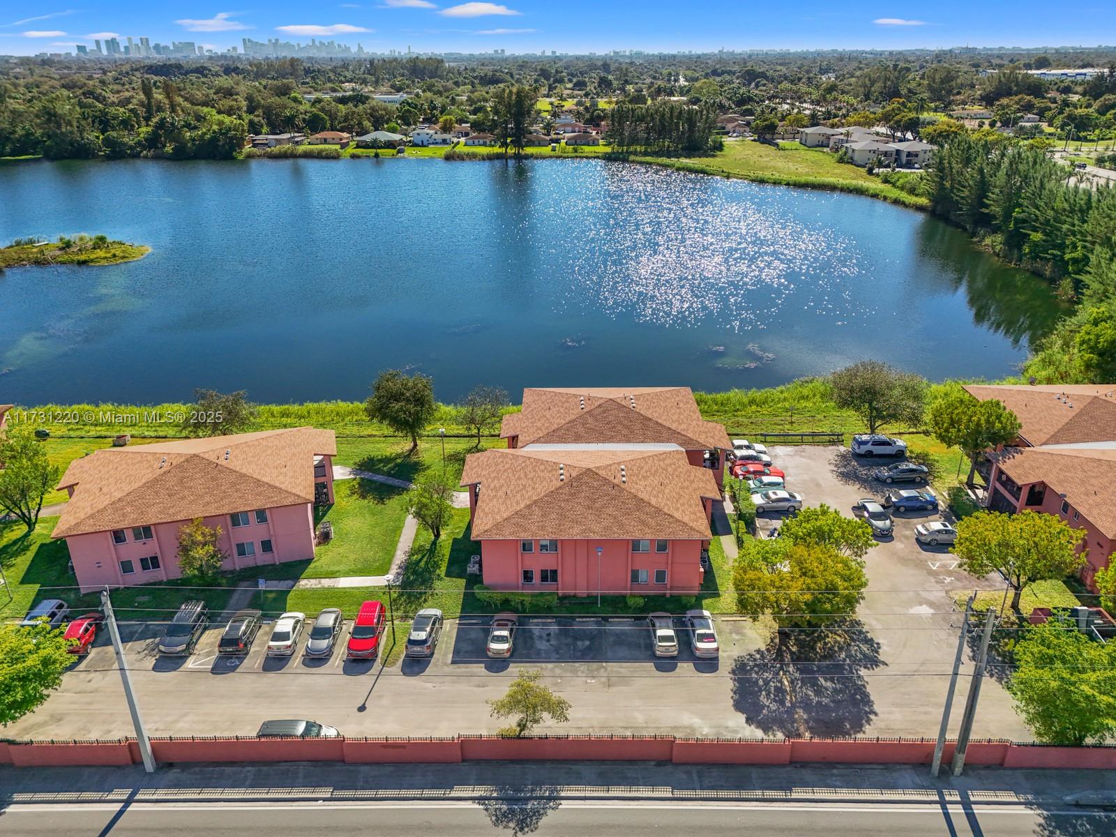 An aerial view shows pink apartment buildings with brown roofs situated beside a large lake — 2020 NW 119th St # 1122, Miami FL 33167, MIAMI, FL