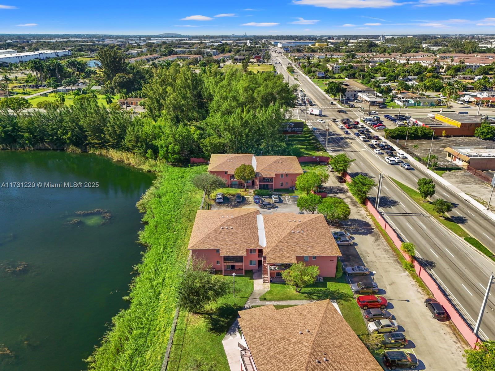 An aerial view shows three multi-story residential buildings situated beside a lake and road — 2020 NW 119th St # 1122, Miami FL 33167, MIAMI, FL