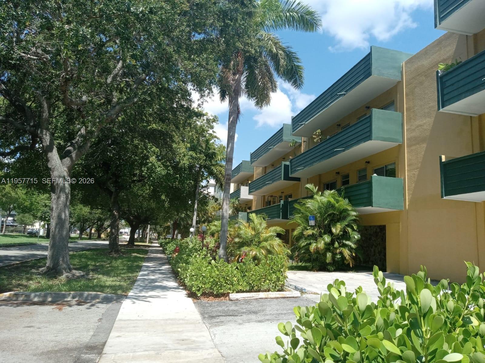 A multi-story apartment building with green balconies stands beside a sidewalk and palm trees — 1975 NE 135th St # 3C, North Miami FL 33181, NORTH MIAMI, FL