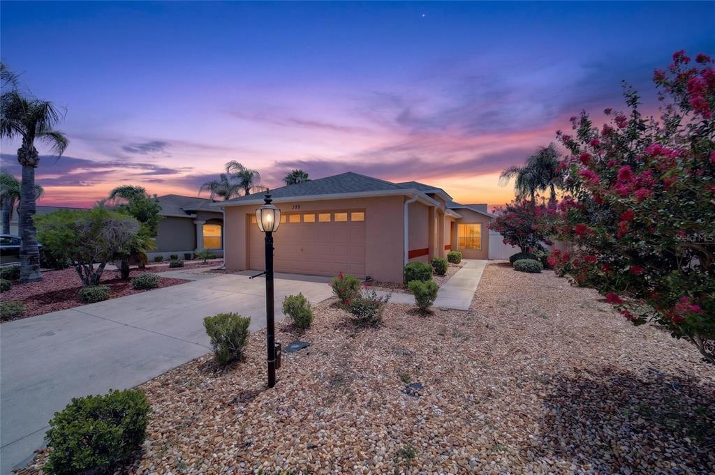 A tan single-story house with a two-car garage and gravel landscaping at sunset — 308 ALMERIA STREET, THE VILLAGES, FL