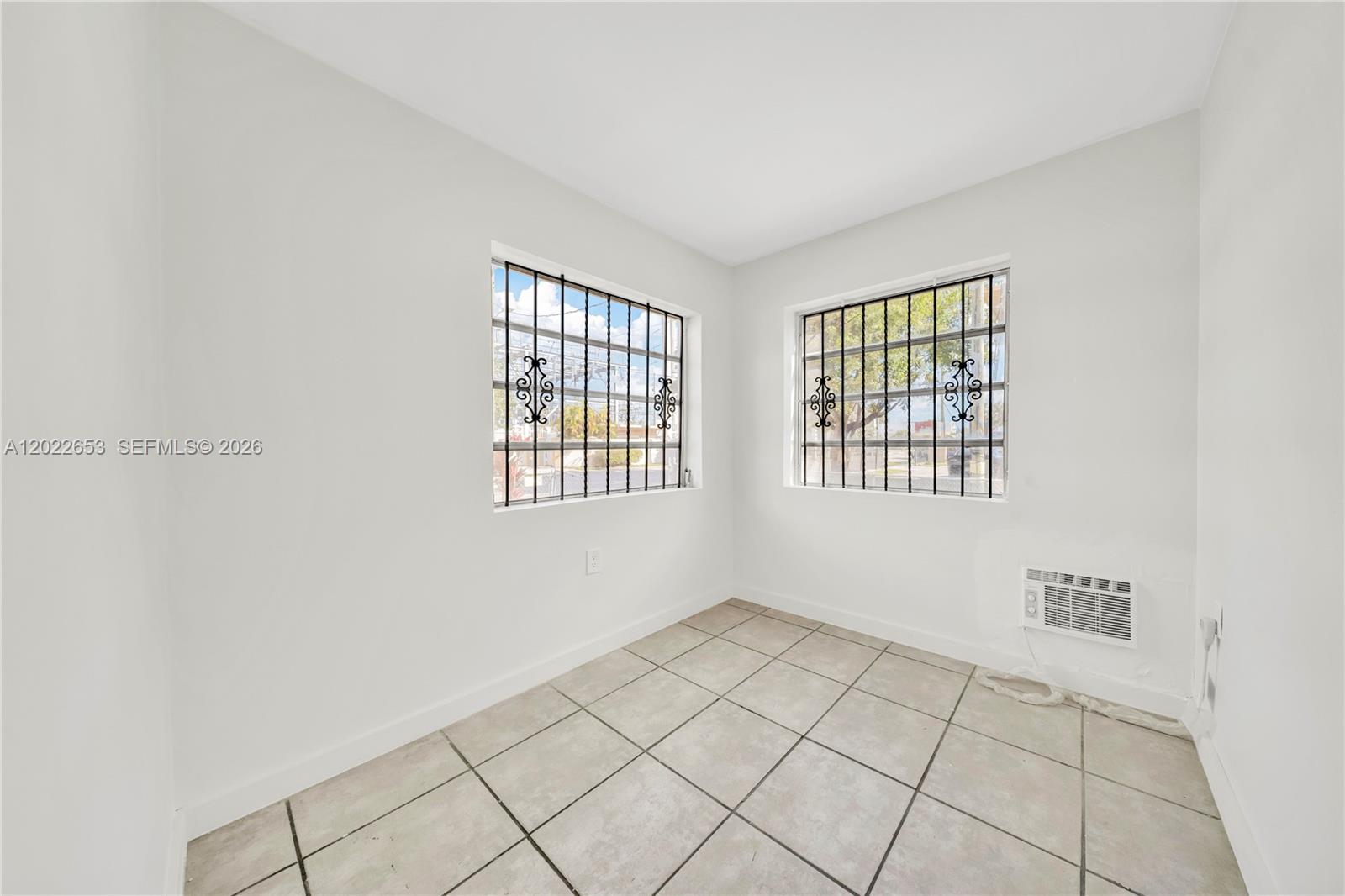 A small, empty room features white walls, tiled floors, two windows with security bars, and an air conditioner — 2744 SW 16th Ter, Miami FL 33145, MIAMI, FL