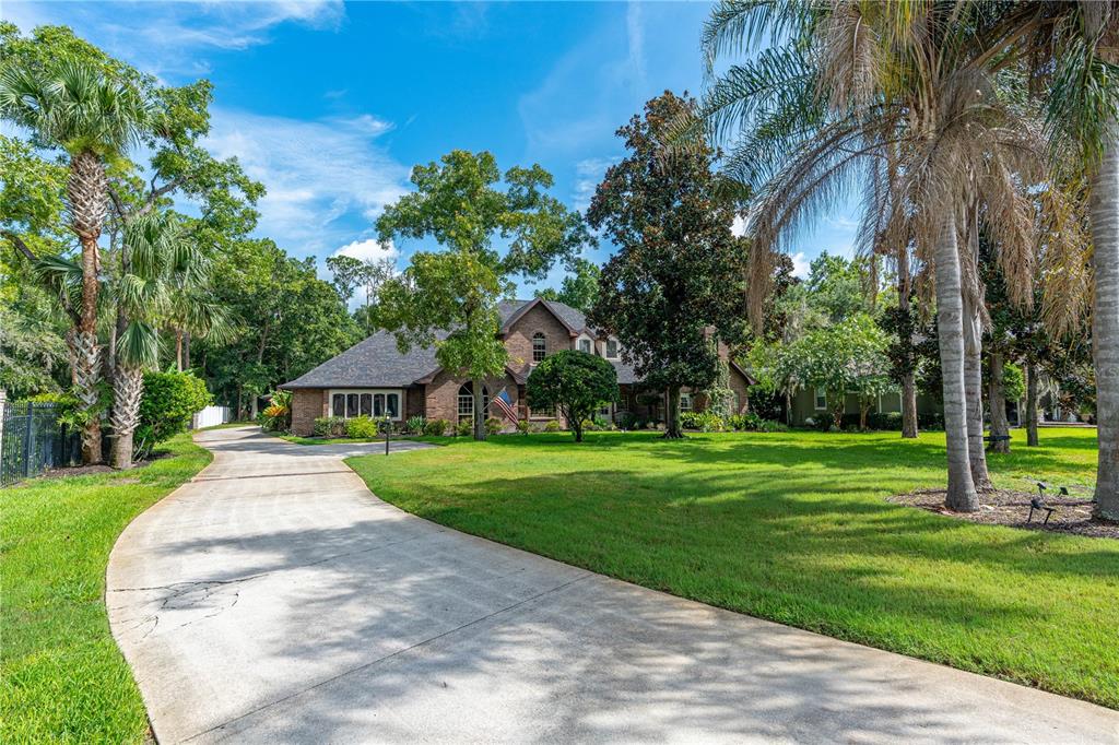 A long concrete driveway leads to a brick house surrounded by trees and green lawn — 73 SHADOW CREEK WAY, ORMOND BEACH, FL