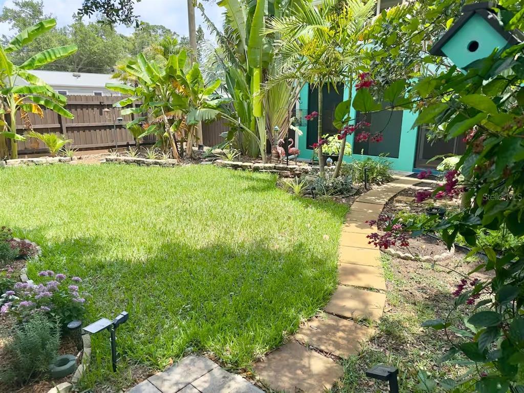A backyard features a stone path, a green lawn, tropical plants, and a wooden fence — 5214 27TH AVENUE S UNIT 2, GULFPORT, FL