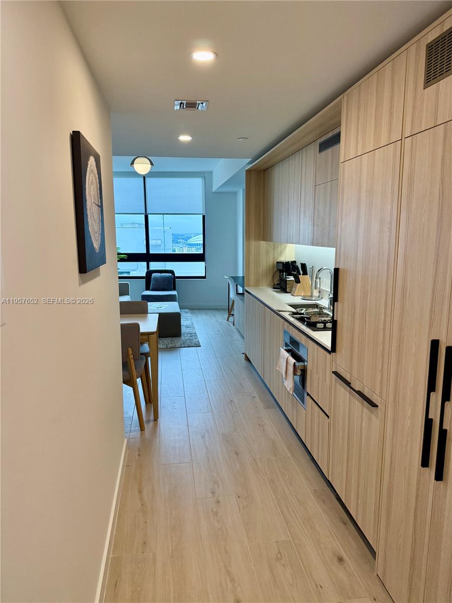 A modern galley kitchen with light wood cabinetry leads to a dining area and window — 225 N Miami Ave # 3406, Miami FL 33128, MIAMI, FL