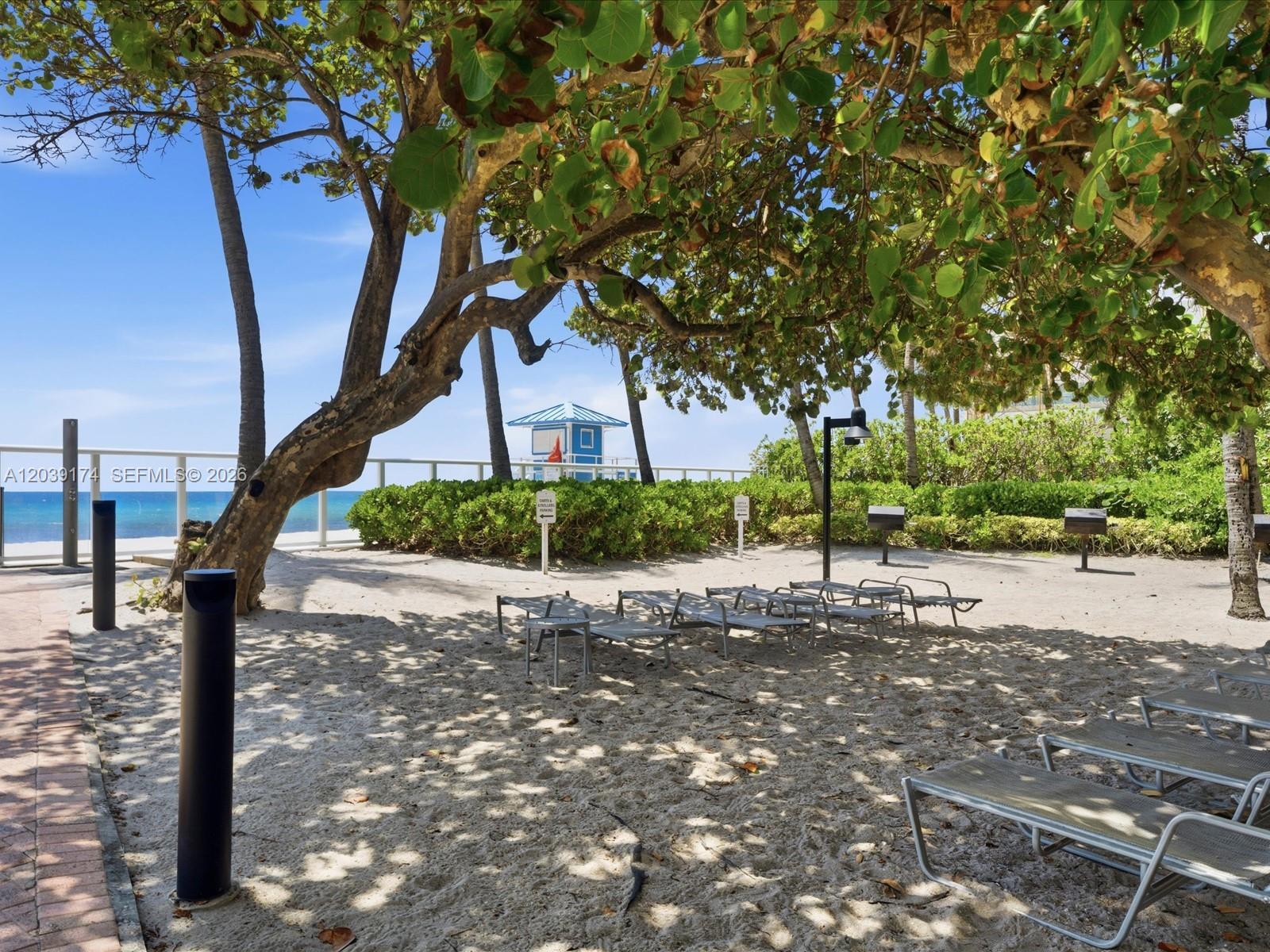 A sandy beach area features lounge chairs under a large tree near a lifeguard tower — 3000 S Ocean Dr # 718, Hollywood FL 33019, HOLLYWOOD, FL