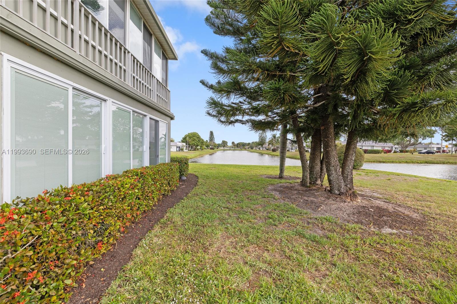 A view of a house exterior with a hedge, pine trees, and a canal — 72 Monaco B # 72, Delray Beach FL 33446, DELRAY BEACH, FL