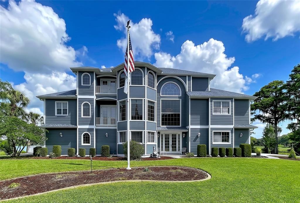 A large, three-story blue house with a central flag pole stands on a grassy lawn — 1360 S WATERVIEW DRIVE, INVERNESS, FL