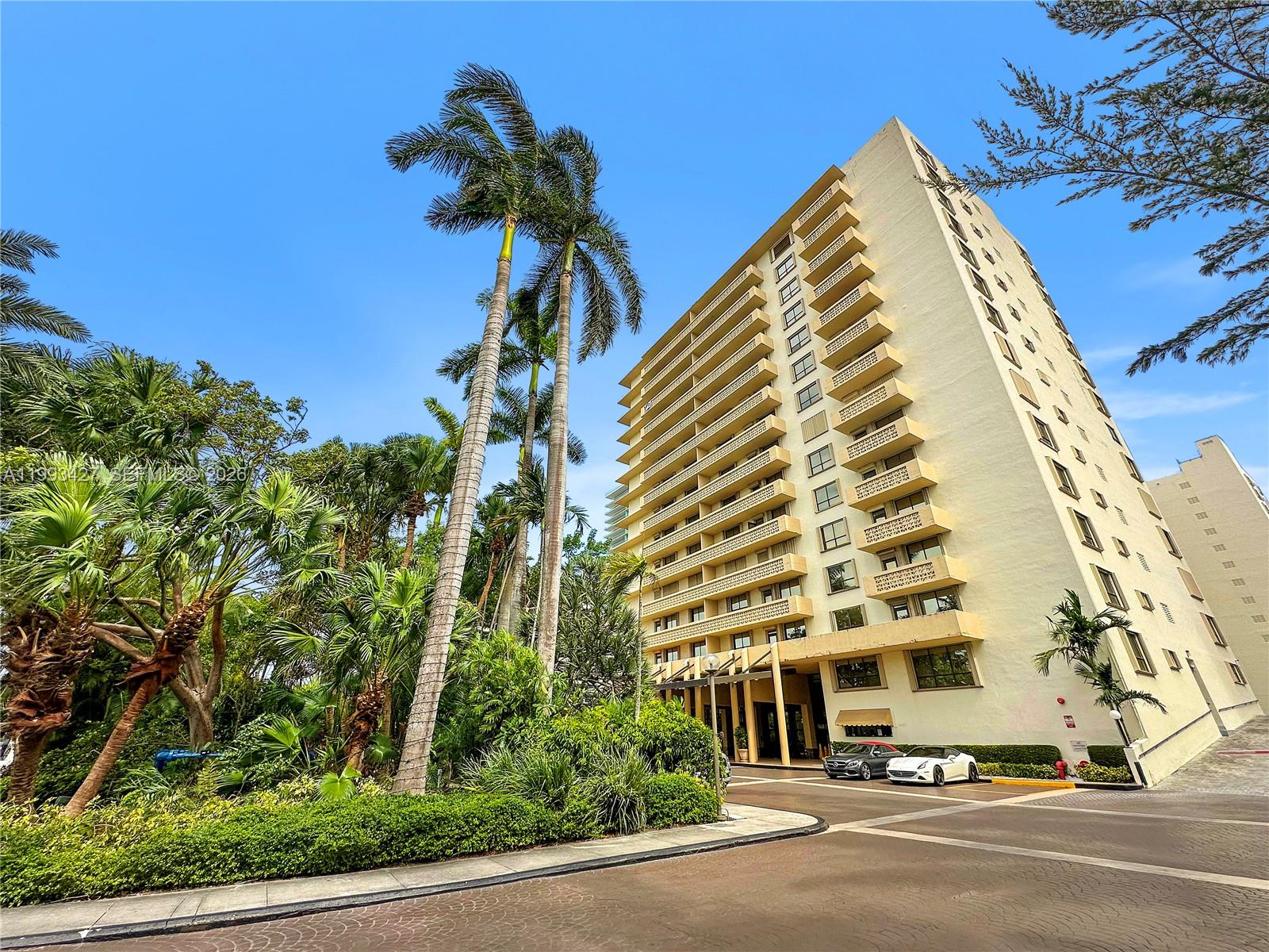 A tall, light-colored apartment building stands behind lush tropical landscaping and a paved driveway — 10185 Collins Ave # 502, Bal Harbour FL 33154, BAL HARBOUR, FL