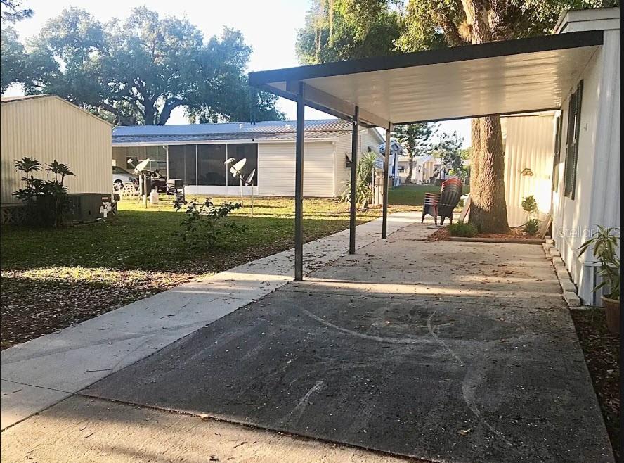 A concrete carport with a metal roof sits beside a mobile home on a grassy lot — 900 AQUA ISLES BOULEVARD UNIT B21, LABELLE, FL