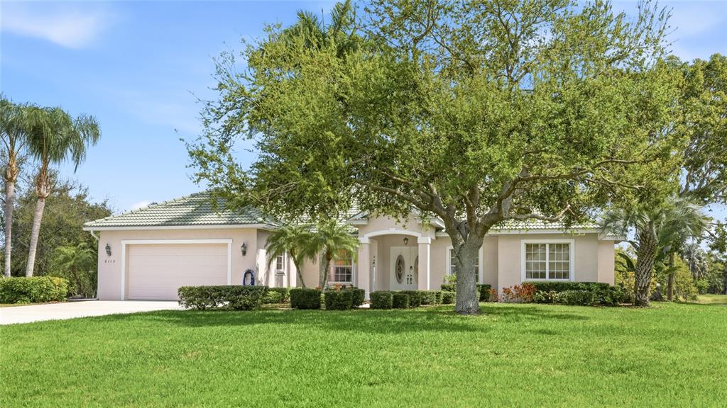 A single-story house with a beige exterior, a two-car garage, and a large front tree — 6115 9TH AVENUE CIRCLE NE, BRADENTON, FL