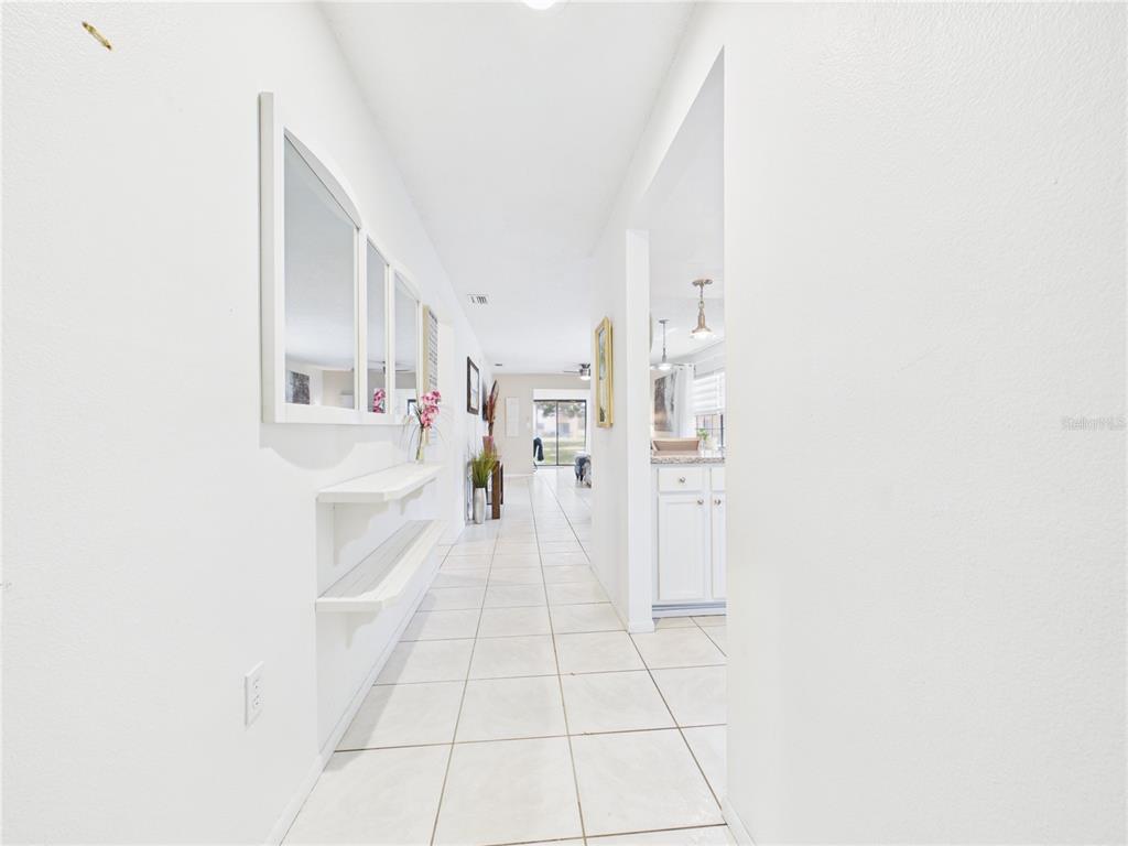 A bright hallway with white walls, tiled floors, and wall-mounted shelves leads toward a kitchen — 13250 RIDGE ROAD UNIT 9-8, LARGO, FL