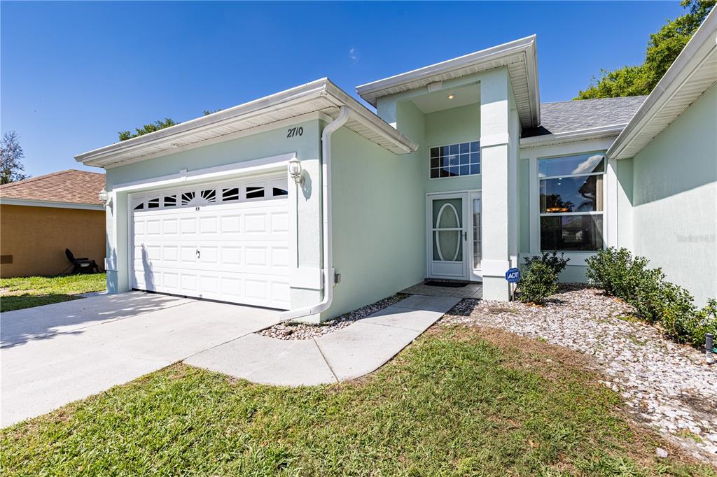 A light green single-story house features a white garage door, a covered entryway, and landscaping — 2710 AUTUMN LANE, EUSTIS, FL