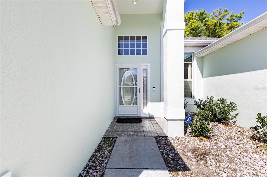 The exterior entrance features a light green stucco wall, a glass storm door, and landscaping — 2710 AUTUMN LANE, EUSTIS, FL