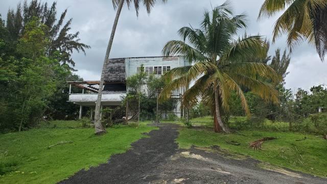 A weathered two-story house with a damaged roof sits on a grassy lot with palm trees — KM. 1.8 PR-678, VEGA ALTA, PR