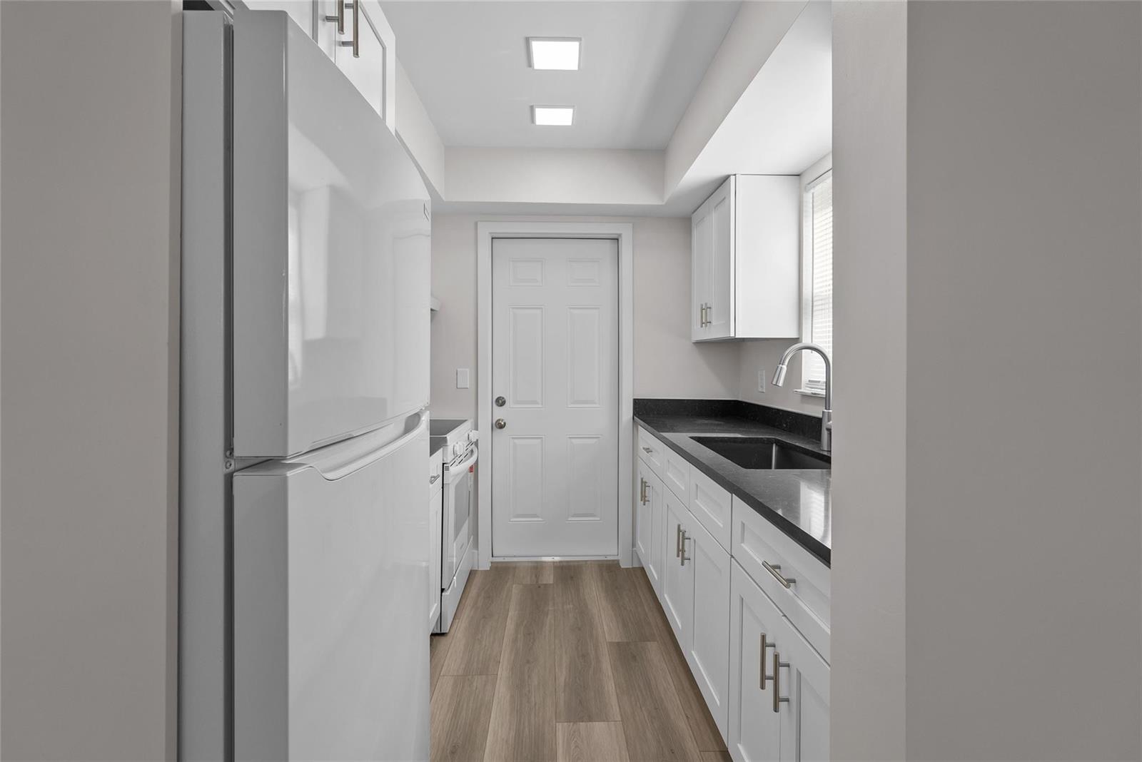 A narrow galley kitchen features white cabinets, dark countertops, stainless appliances, and wood-look flooring — 1150 E Sample Road # 202, Pompano Beach FL 33064, POMPANO BEACH, FL