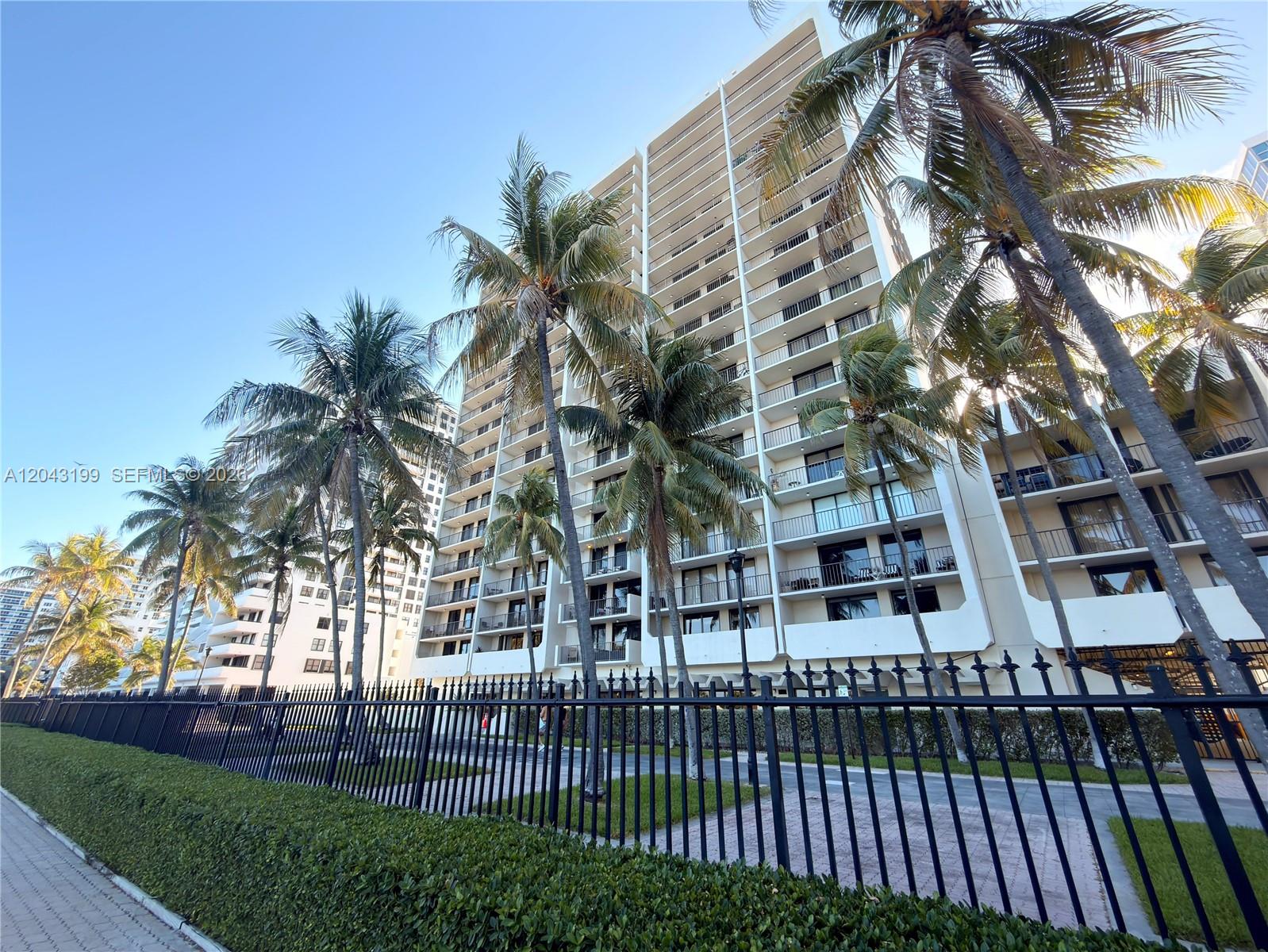 A tall residential building stands behind a black metal fence and several palm trees — 2625 Collins Ave # 215, Miami Beach FL 33140, MIAMI BEACH, FL