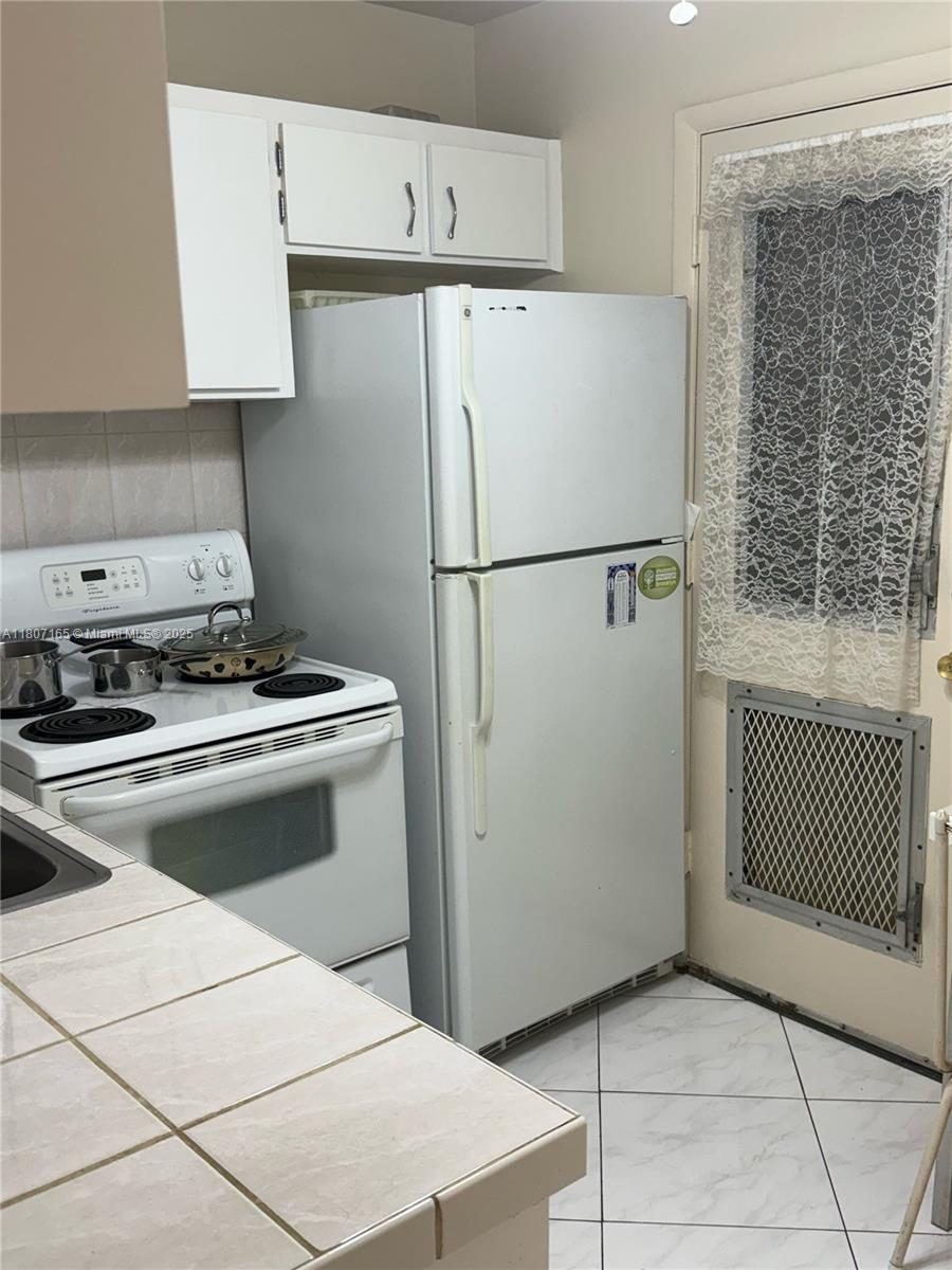 A kitchen features white appliances, tiled countertops, white cabinets, and a door with a lace curtain — 1395 NE 167th St # 203, Miami FL 33162, MIAMI, FL