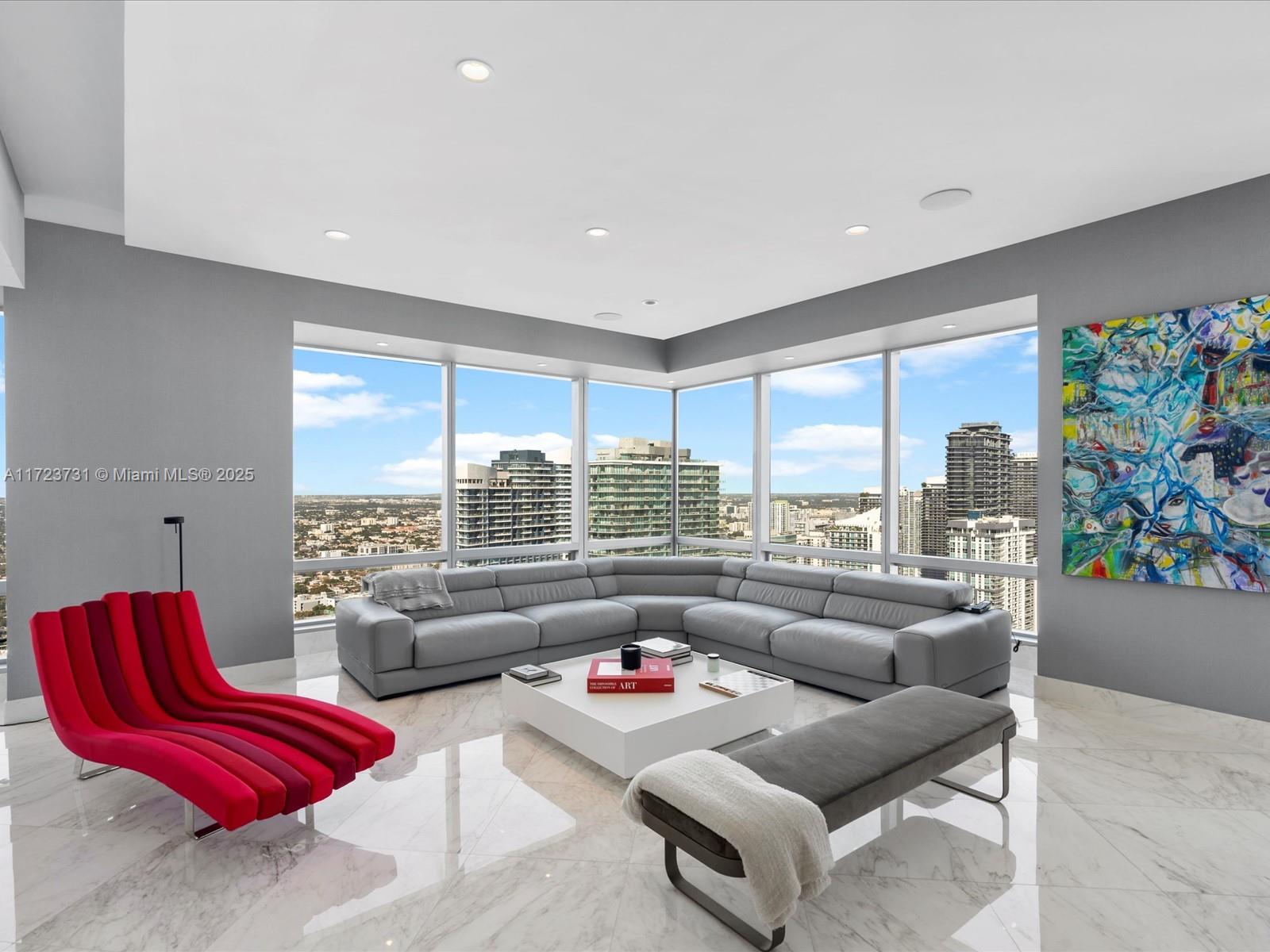 A modern living room features a grey sectional, red lounge chair, and floor-to-ceiling city views — 1425 Brickell Ave, Miami FL 33131, MIAMI, FL