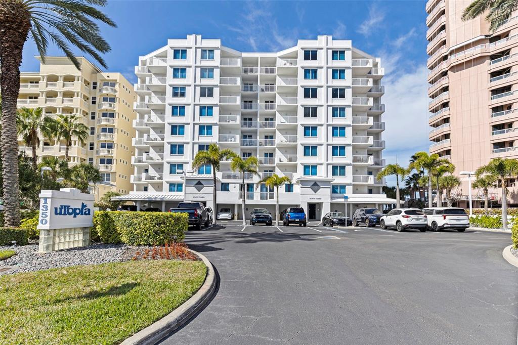 A multi-story white residential building with balconies stands behind a paved parking lot — 1350 GULF BOULEVARD UNIT 704, CLEARWATER BEACH, FL