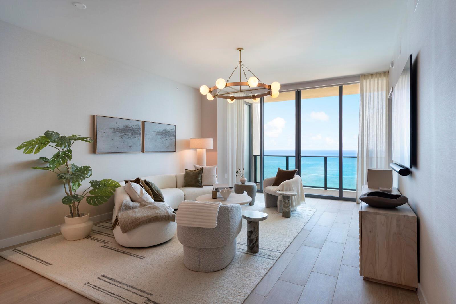 A modern living room features a curved sofa, neutral furniture, and floor-to-ceiling windows overlooking the ocean — 900 N Ocean Boulevard, Pompano Beach FL 33062, POMPANO BEACH, FL