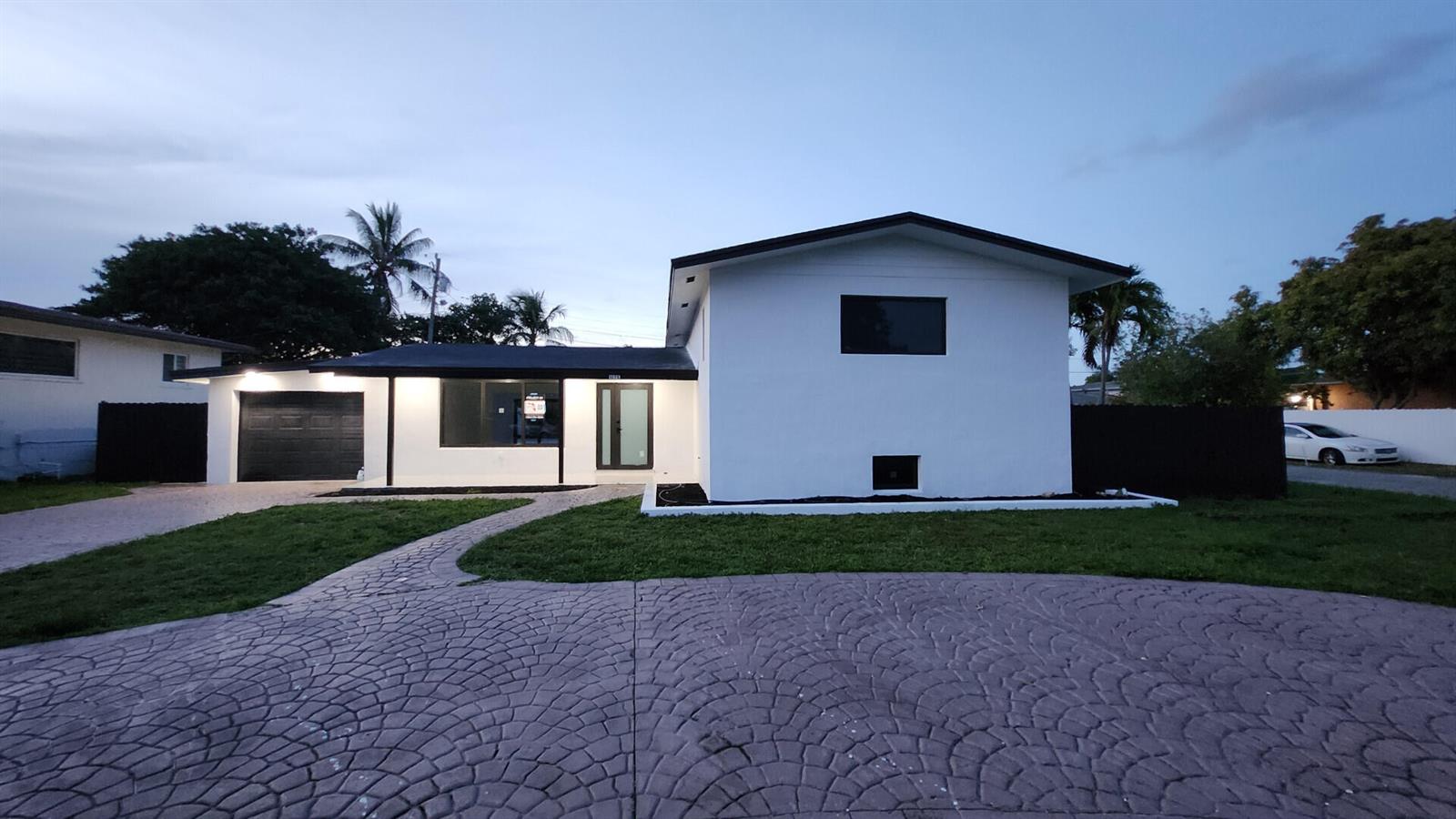 A white single-story house with a black roof, garage, and patterned concrete driveway at dusk — 1275 NE 200th Terrace, Miami FL 33179, MIAMI, FL