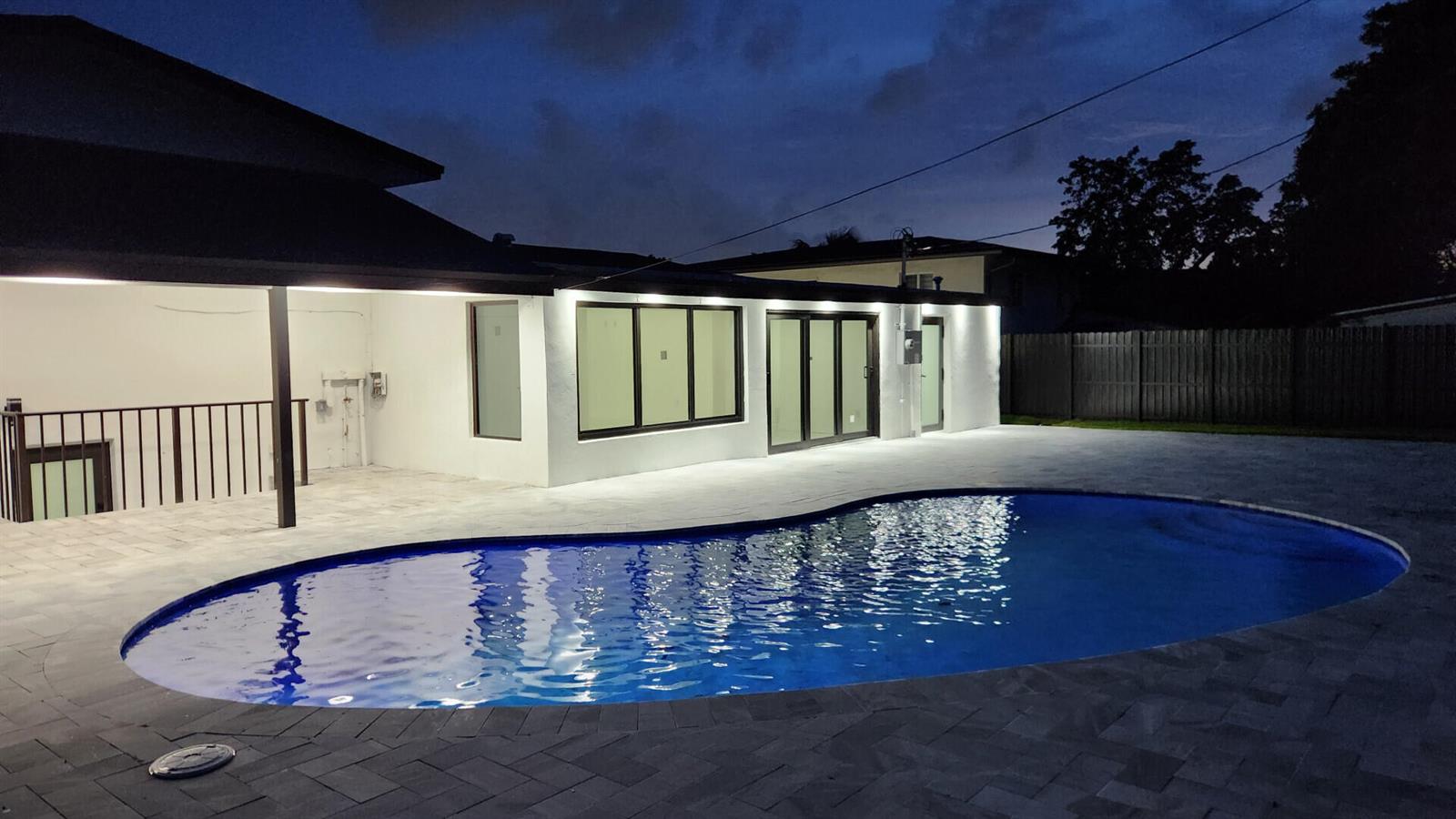 A backyard swimming pool sits next to a white house with modern windows at dusk — 1275 NE 200th Terrace, Miami FL 33179, MIAMI, FL
