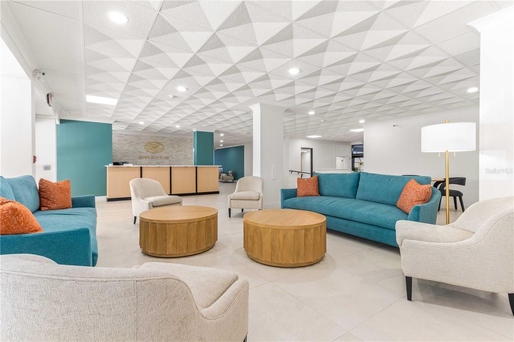 A modern lobby features teal sofas, light-colored armchairs, round wooden coffee tables, and a textured ceiling — 1380 GULF BOULEVARD UNIT 307, CLEARWATER BEACH, FL