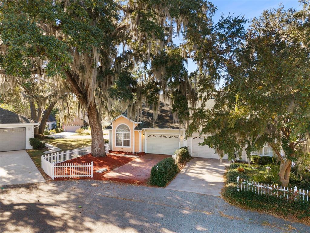 A tan house with a brick driveway and white picket fence sits under large trees — 10804 NW 65TH WAY, ALACHUA, FL