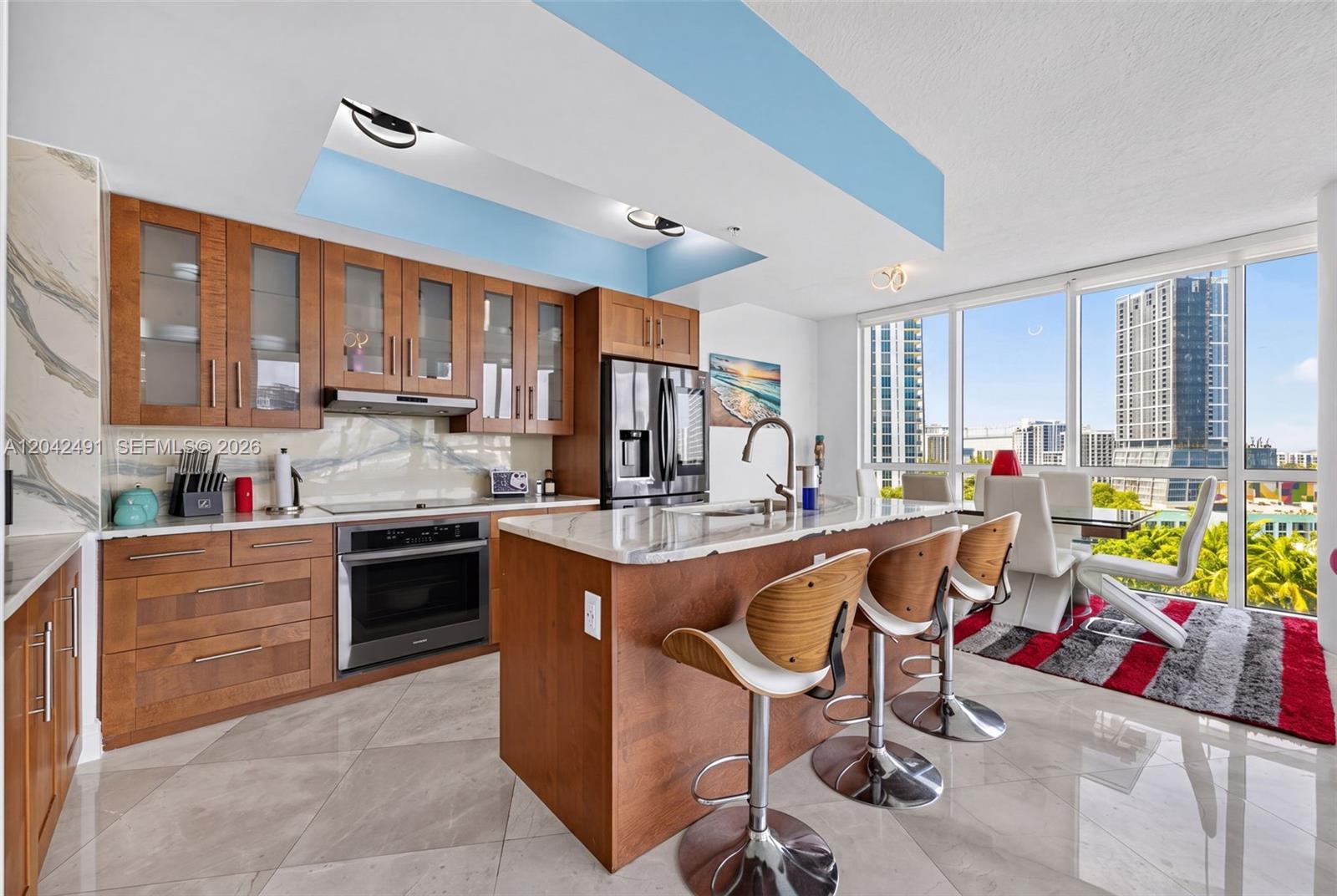 A modern kitchen features wood cabinetry, a marble-topped island, and floor-to-ceiling windows with city views — 480 NE 30th St # 1807, Miami FL 33137, MIAMI, FL