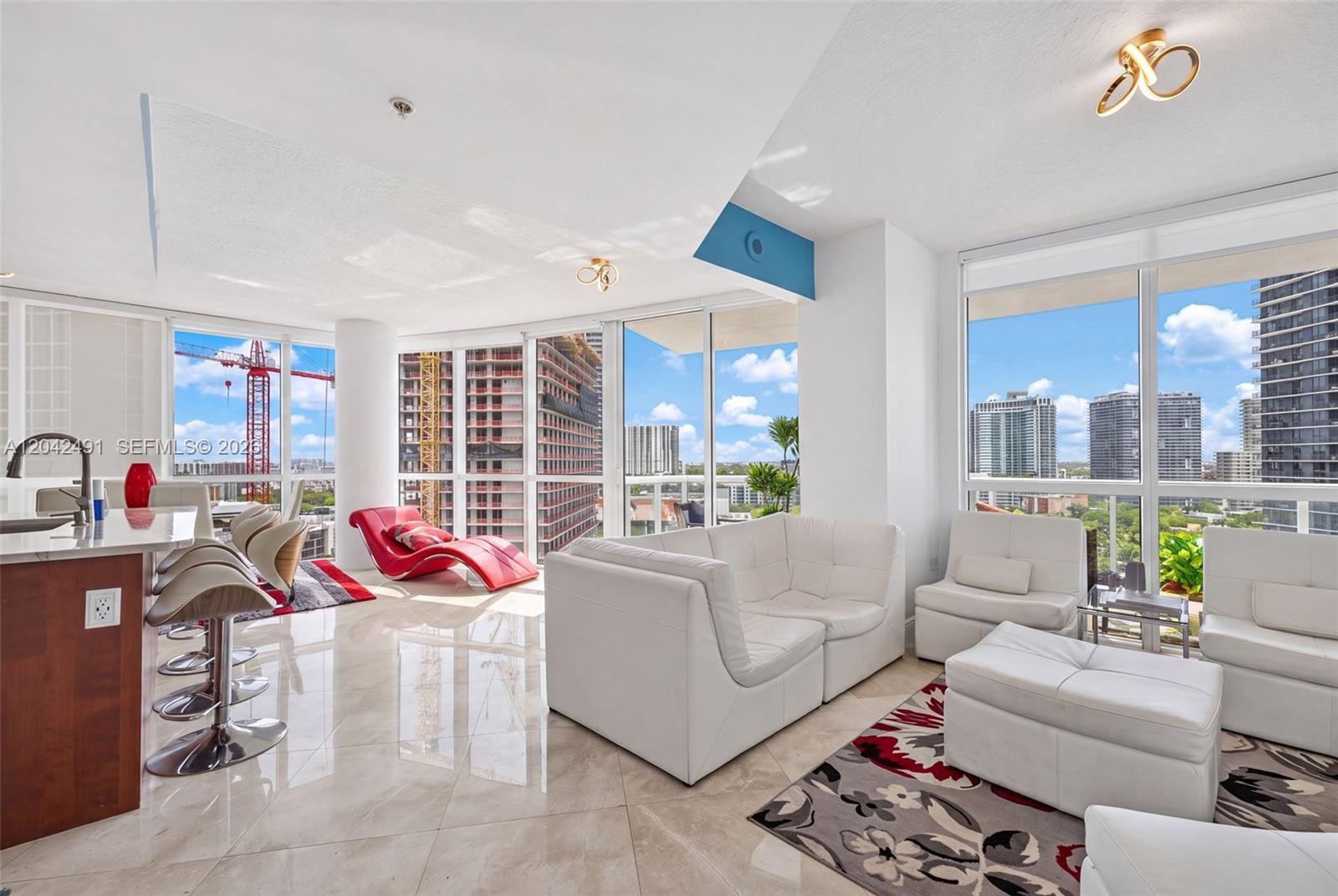 A modern living room features white leather furniture, tile floors, and floor-to-ceiling windows with city views — 480 NE 30th St # 1807, Miami FL 33137, MIAMI, FL