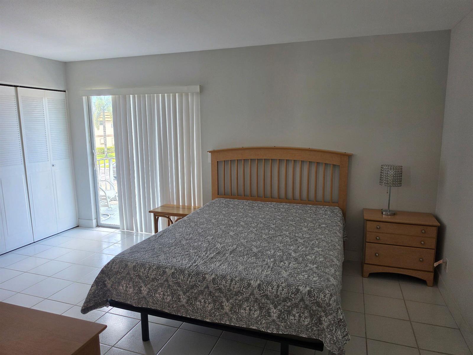A bedroom with a wooden bed frame, a nightstand, tiled floors, and a sliding glass door — 79 Brittany # B, Delray Beach FL 33446, DELRAY BEACH, FL