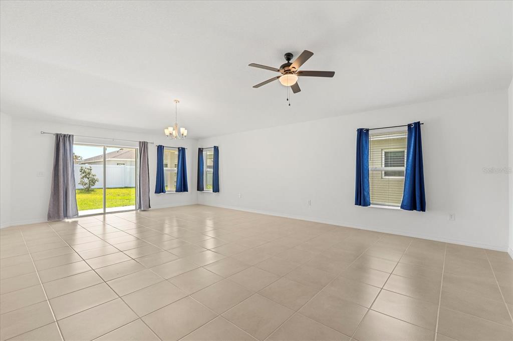A spacious, white-walled living room features light tile flooring, a ceiling fan, and blue curtains — 1429 MATTIE POINTE LANE, AUBURNDALE, FL