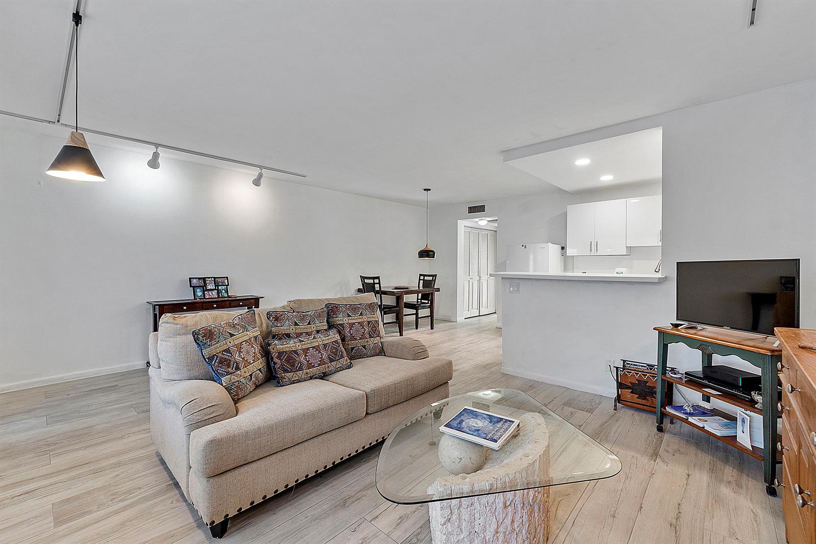 A living room with a beige sofa, glass coffee table, and an open kitchen area — 1012 Newport G, Deerfield Beach FL 33442, DEERFIELD BEACH, FL