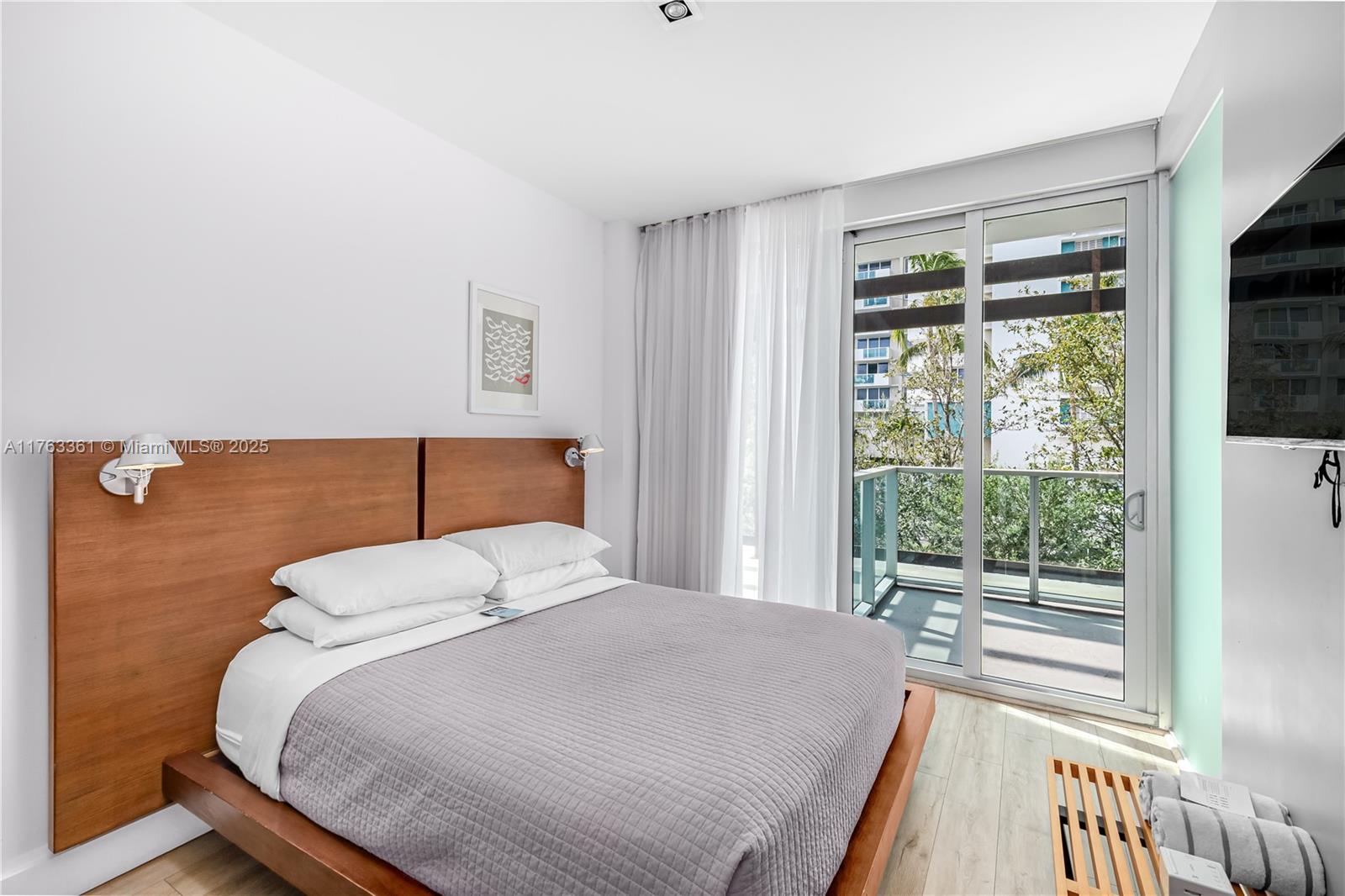 A bedroom features a wooden bed frame, white bedding, and a sliding glass door to a balcony — 1215 West Ave # 203, Miami Beach FL 33139, MIAMI BEACH, FL