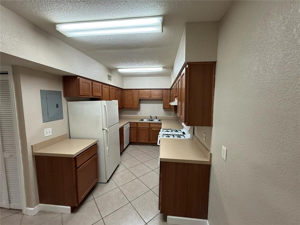 A kitchen features wood cabinets, beige countertops, white appliances, and light-colored tile flooring — 1355 DREW STREET UNIT 101, CLEARWATER, FL