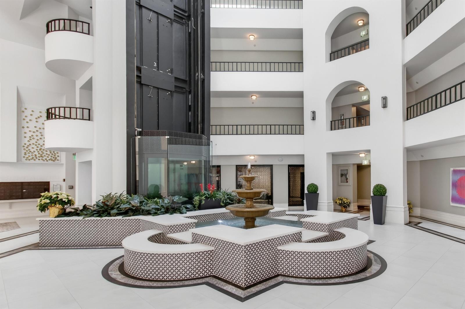 A multi-story atrium features a central tiered fountain, white tiled floors, and a glass elevator — 140 SE 5th Avenue # 448, Boca Raton FL 33432, BOCA RATON, FL