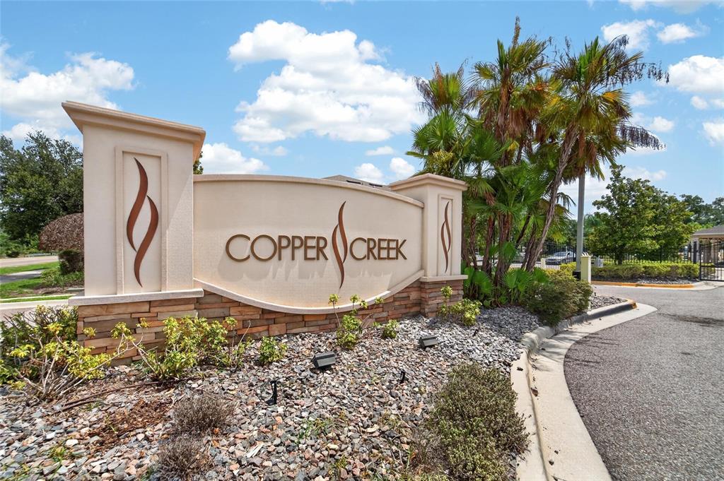 A beige stucco monument sign for Copper Creek stands near a paved road and palm trees — 12925 SHADY FERN LANE, GIBSONTON, FL