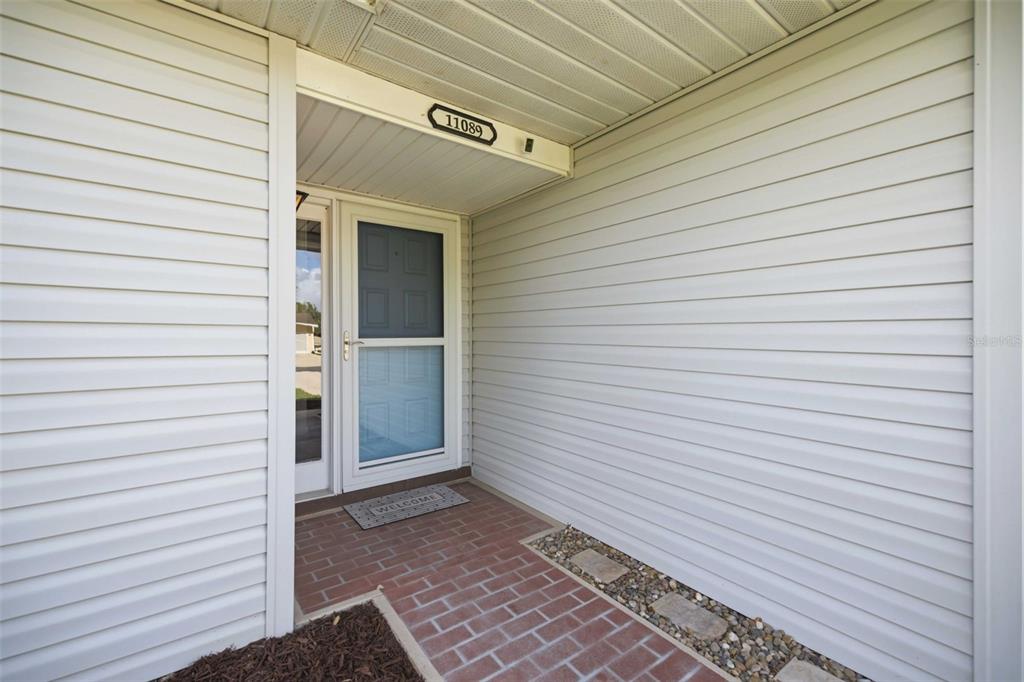 A covered entryway features white vinyl siding, a brick walkway, and a glass storm door — 11089 SW 78TH COURT, OCALA, FL