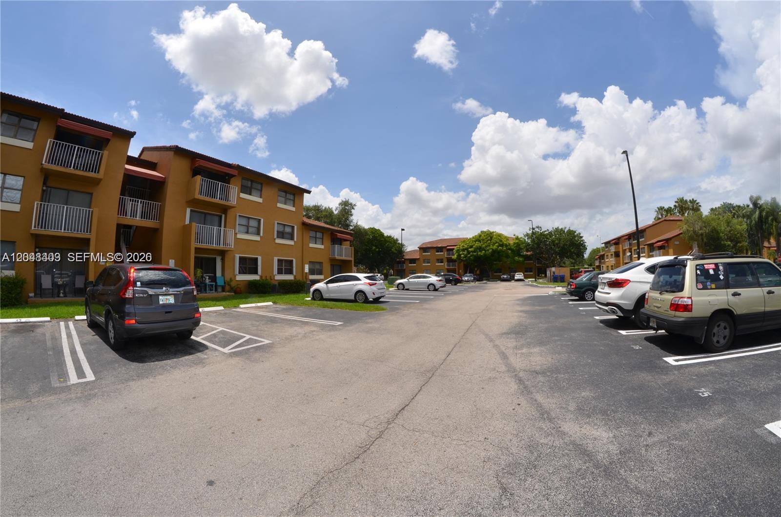 An outdoor parking lot sits in front of multi-story apartment buildings under a blue sky — 15630 SW 80th St # 210, Miami FL 33193, MIAMI, FL
