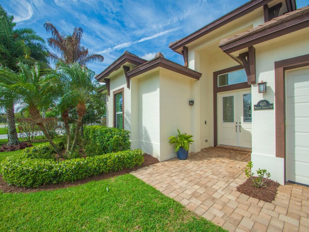 A paved walkway leads to the front entrance of a house with cream-colored stucco walls — 445 Wingate Terrace SW, Vero Beach FL 32968, VERO BEACH, FL