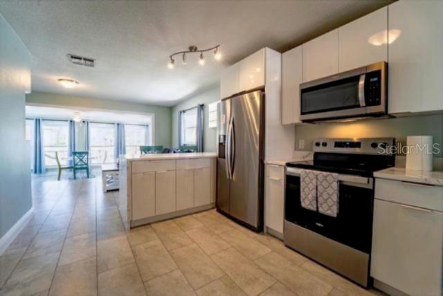 A modern kitchen with white cabinets, stainless steel appliances, and tile flooring opens to a dining area — 4825 FLORAMAR TERRACE, NEW PORT RICHEY, FL