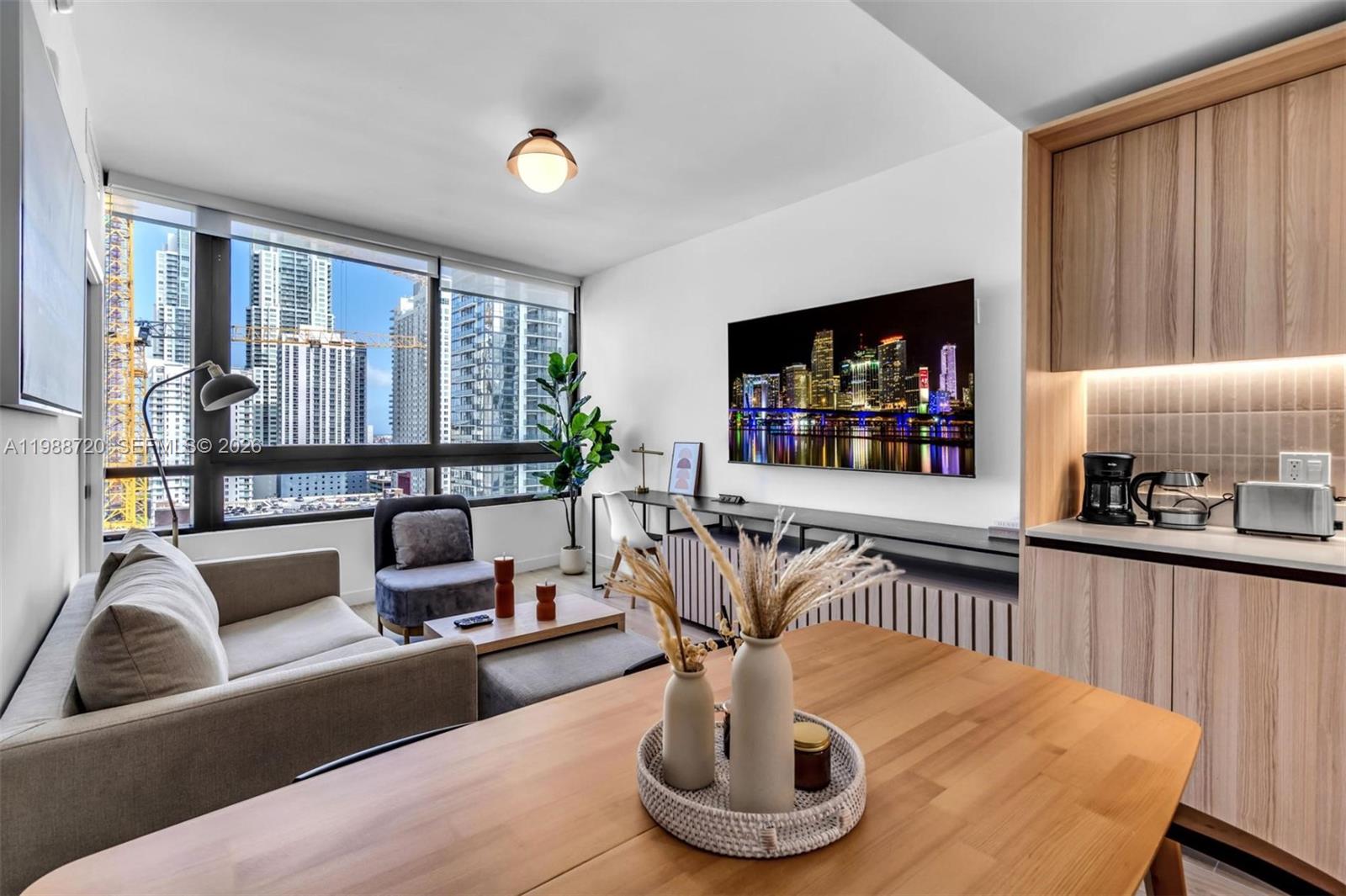 A modern living room with a sofa, dining table, and large windows overlooking city skyscrapers — 225 N Miami Ave # 1701, Miami FL 33131, MIAMI, FL