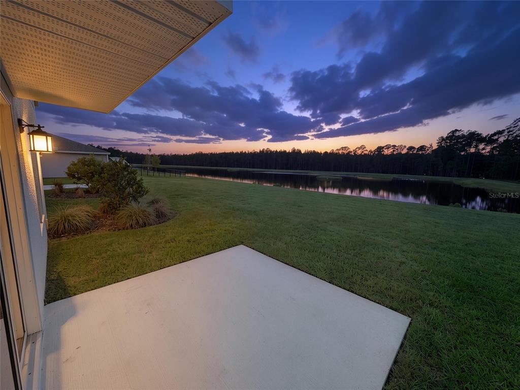 A concrete patio overlooks a grassy lawn, a pond, and a treeline at sunset — 1185 SAND TRAP COURT, DAYTONA BEACH, FL