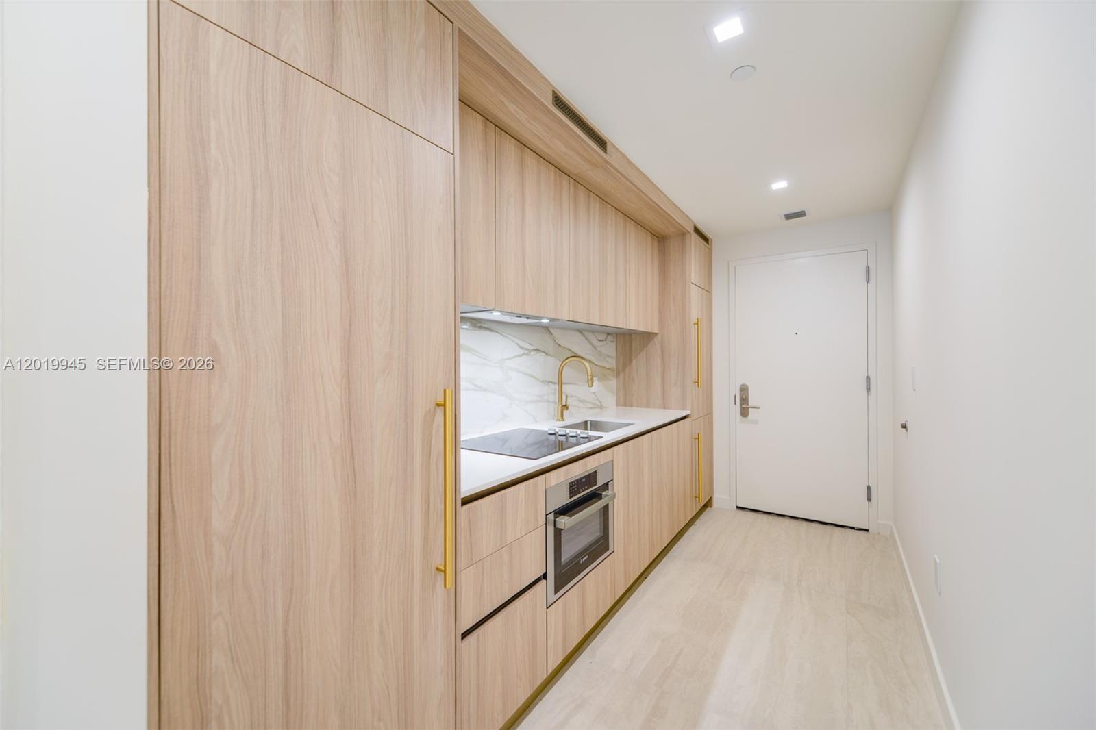 A modern galley kitchen features light wood cabinetry, gold hardware, and a marble backsplash — 77 5th St # 2604, Miami FL 33131, MIAMI, FL