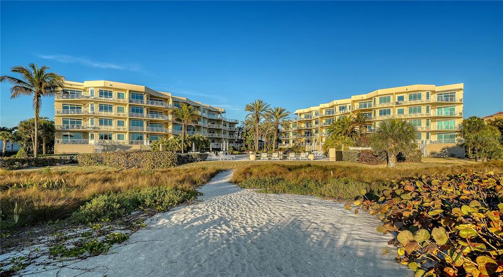 A sandy path leads toward two multi-story apartment buildings surrounded by palm trees and vegetation — 2165 GULF OF MEXICO DRIVE UNIT 115, LONGBOAT KEY, FL