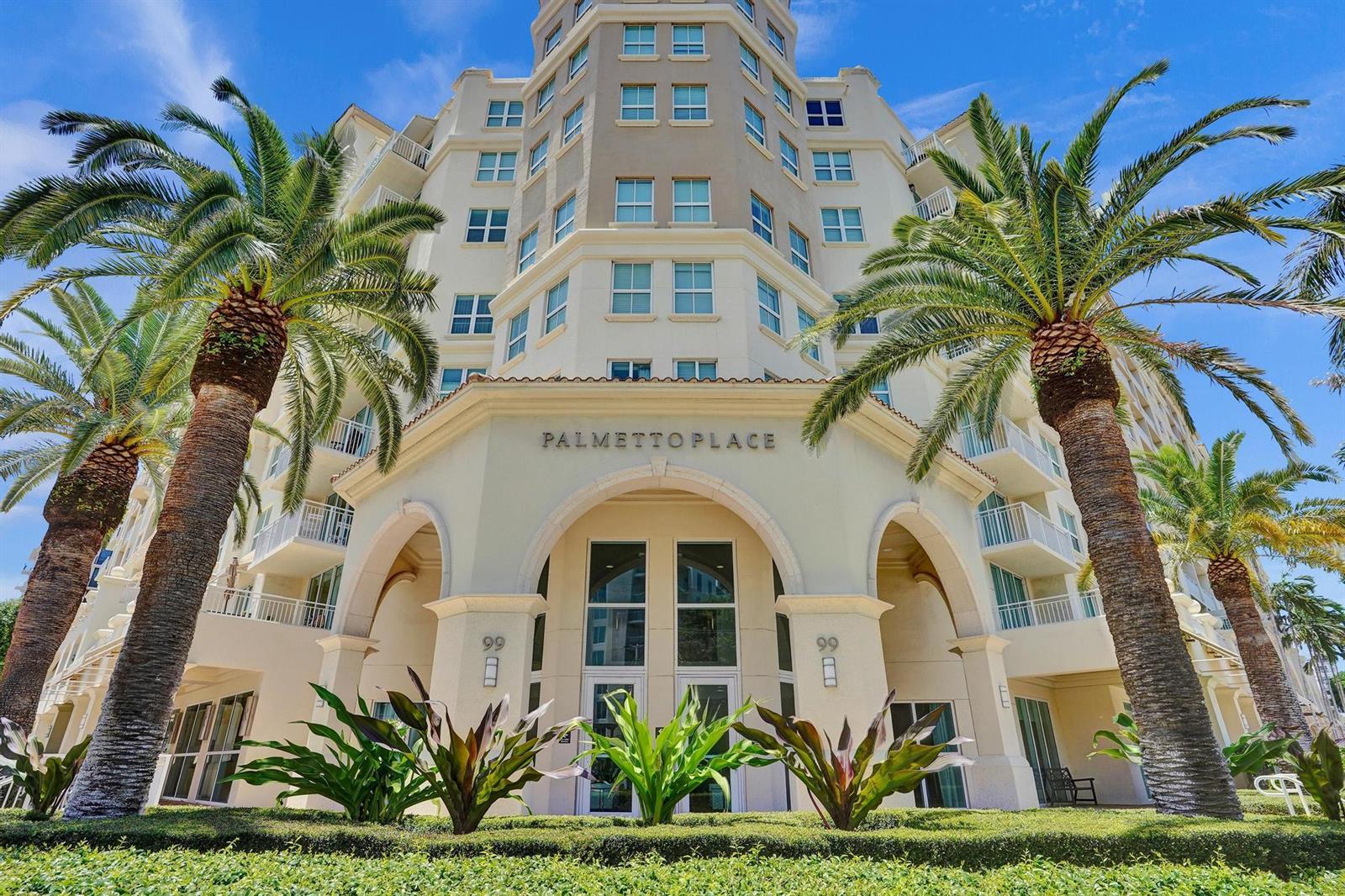 The exterior of a multi-story apartment building features palm trees and a prominent arched entrance — 99 SE Mizner Boulevard # 40, Boca Raton FL 33432, BOCA RATON, FL