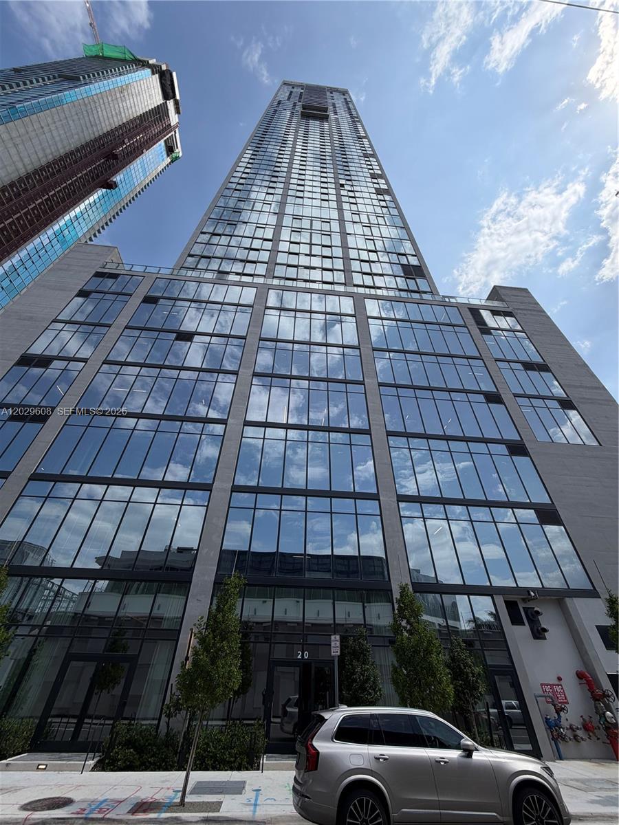 A low-angle exterior view of a modern glass skyscraper with a silver SUV parked in front — 20 NE 11TH ST # 3306B, Miami FL 33132, MIAMI, FL