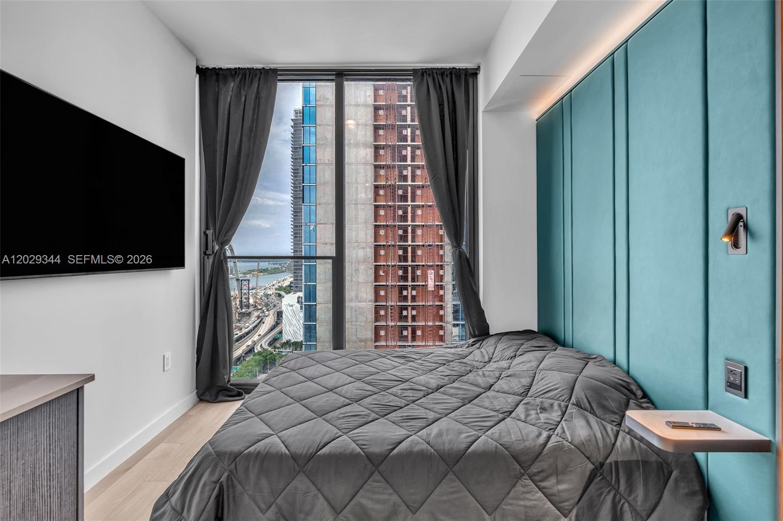 A bedroom features a teal upholstered headboard, a quilted bed, and a floor-to-ceiling window — 20 NE 11th St # 2606A, Miami FL 33132, MIAMI, FL