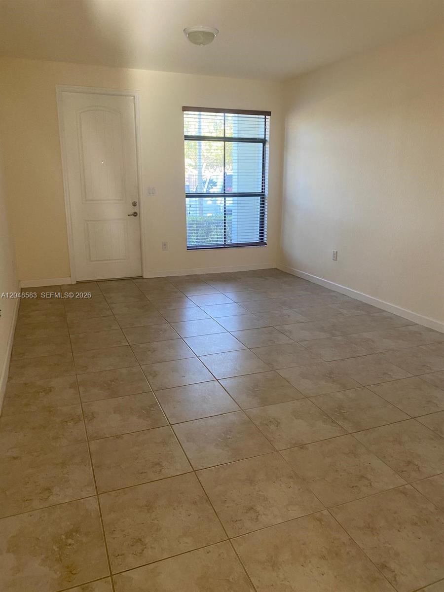 An empty room features beige tile flooring, a white door, and a window with blinds — MIAMI, FL