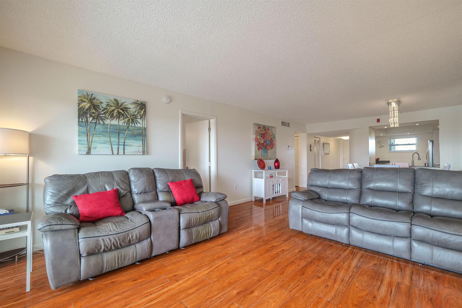 A living room features two gray leather sofas, wood-look tile floors, and palm tree artwork — 3930 Inverrary Boulevard # d, Lauderhill FL 33319, LAUDERHILL, FL